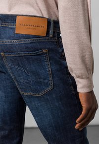 Denim jeans in dark blue with a faded finish. Features a tan leather patch labeled "BALDESSARINI JEANS" and contrasting yellow stitching.