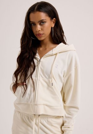 Frau mit langen dunklen Haaren, die große Creolen und einen cremefarbenen Kapuzenpullover mit Reißverschluss sowie passende Jogginghosen mit Kordelzug trägt und mit einer Hand in der Tasche posiert.