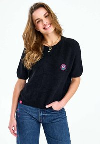 Mlada žena koja se smiješi, ima labavo valovitu kosu i nosi crni džemper kratkih rukava s cvjetnim aplikacijama te plave traperice, ruka joj je u džepu.