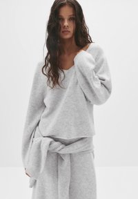 Pulover oversized, gri deschis, din material moale, cu decolteu larg și mâneci lungi, asociat cu un detaliu legat corespunzător la talie.