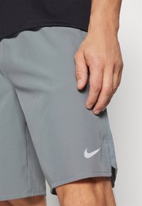 Graue Sportshorts aus leichtem Material, mit einem seitlichen Schlitz und einem weißen Nike-Logo am linken Saum. Glatte Textur.