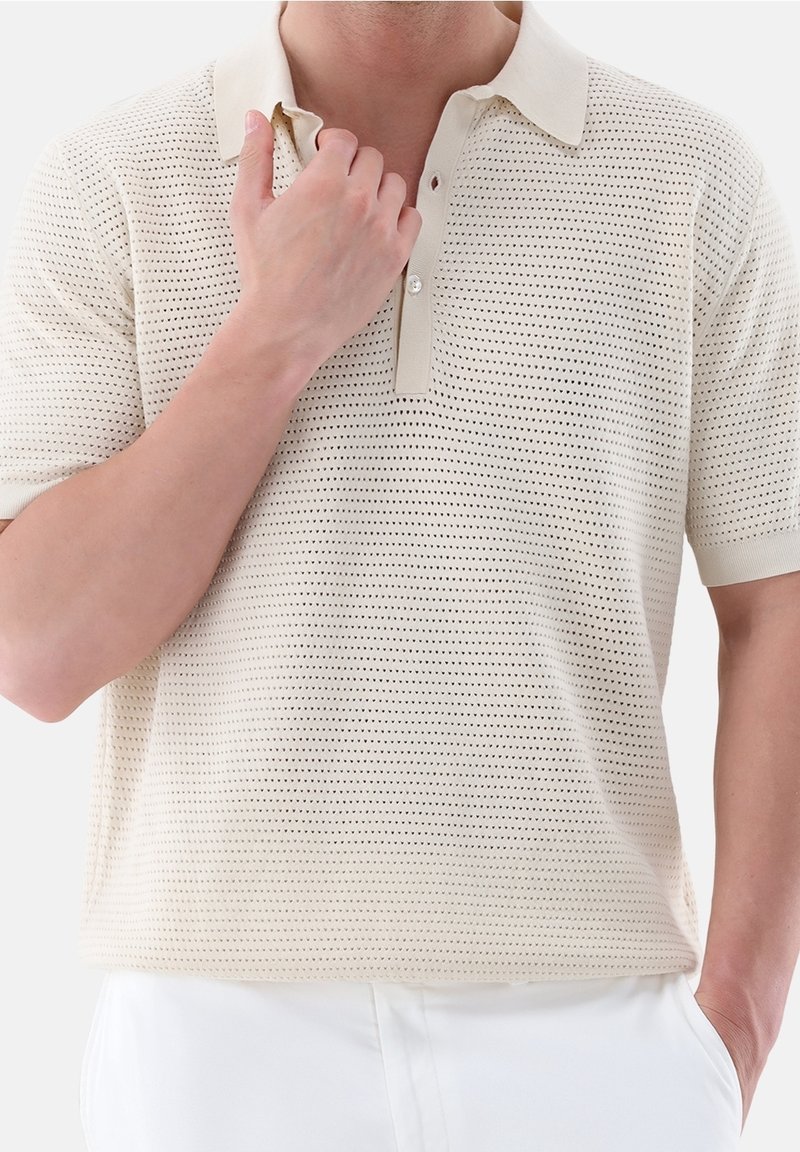 Homme portant un polo en maille beige à manches courtes avec de petits trous, une main ajustant le col, l'autre main dans la poche d'un pantalon blanc.