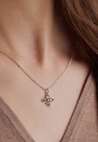 Collana in argento con pendente floreale decorato con gemme arancioni e trasparenti, dettagli intricati e finitura lucida, appoggiata su una maglia marrone.