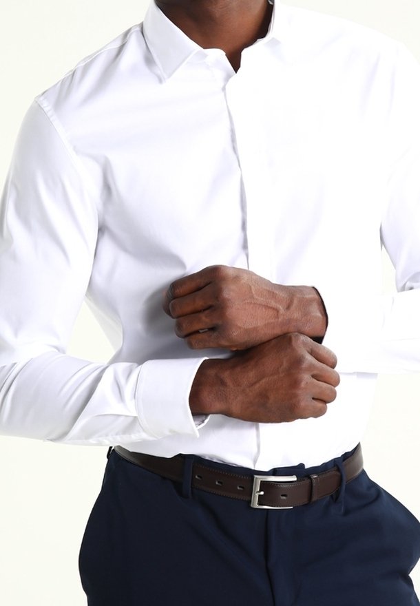Camisa blanca de manga larga con cuello, combinada con pantalones oscuros y un cinturón de cuero marrón, que muestra una tela suave y un diseño ajustado.