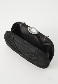 Borsa a mano nera con un esterno strutturato, caratterizzata da un design a torsione e una grande chiusura con strass. Interno foderato in morbido tessuto nero.