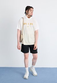 White and yellow striped Nike T-shirt with "AIR" logo, black shorts with side stripes, white socks, and bulky white sneakers.