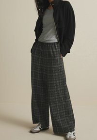 Pantalon large noir texturé avec un motif à carreaux blanc, taille élastique, associé à un t-shirt gris et une veste noire. Chaussures grises et noires.
