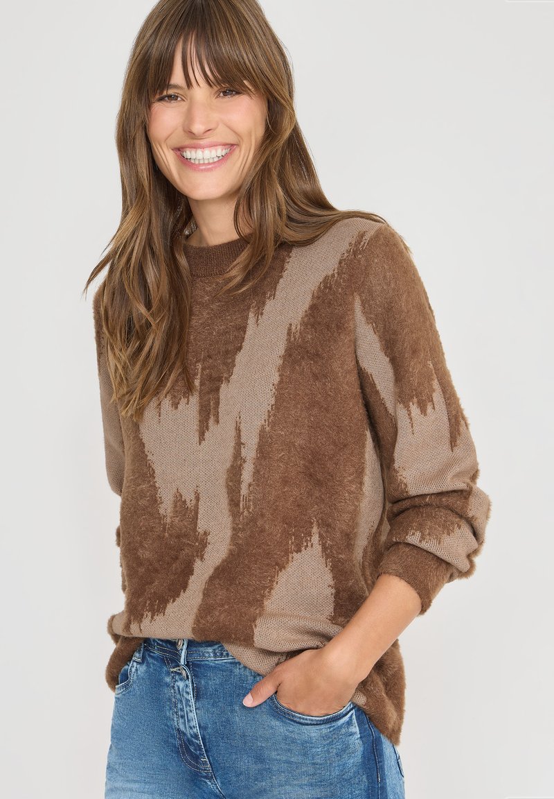 Glimlachende vrouw met lang bruin haar, gekleed in een bruin patroontje fuzzy trui en blauwe jeans, met één hand in haar zak tegen een witte achtergrond.