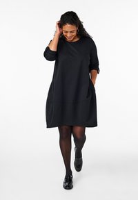 Vestido negro de corte amplio con mangas de tres cuartos, dobladillo curvado y bolsillos laterales. Tejido suave con una textura blanda. Medias negras y mocasines.