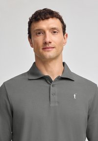 Grå långärmad polo med strukturerad tyg, tre knappar vid kragen och en liten vit golfare-logo på vänster bröst.