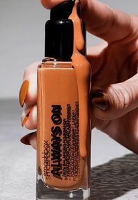 Flacon pompe en verre transparent avec un couvercle noir. Le liquide est d'une teinte orange chaud, gouttant du bec verseur. Le texte sur l'étiquette est en gras et noir.