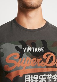 Šedé bavlněné tričko s maskáčovým potiskem, oranžovým a bílým grafickým textem, krátkými rukávy a kulatým výstřihem.