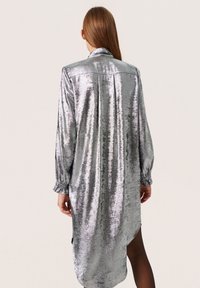 Vestido plateado metálico de manga larga con tejido texturizado, cuello con solapas, puños ajustados y un dobladillo asimétrico.