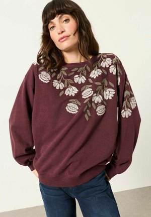 Woman wearing a maroon sweater with white and green floral embroidery on shoulders and sleeves, paired with blue jeans, standing against a plain background.
