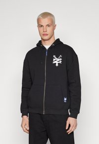 Svart hettegenser med glidelås i bomullsblanding, med hvit grafisk design på brystet, frontlommer og snor i hetten.
