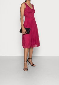 Robe fuchsia avec corsage en dentelle et jupe plissée, associée à une pochette noire et des talons à lanières. Texture lisse et encolure en V.