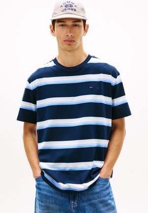 Ung mand iført en marineblå og hvidstribet t-shirt, blå jeans og en beige Tommy Jeans kasket, hænderne i lommerne, neutral ansigtsudtryk.