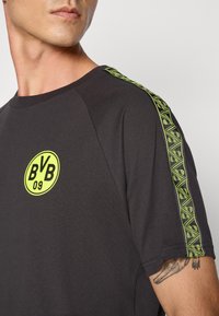 Sort kortærmet t-shirt med rund halsudskæring, prydet med et gult cirkulært logo på brystet og mønstrede grønne striber langs skuldrene.