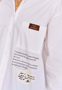 Biały koszula z guzikiem z brązową metką NAOKO na kieszeni. Zawiera nadrukowany tekst oraz notatkę na tkaninie. Miękka, gładka faktura.