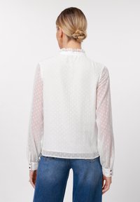 White blouse with sheer, dotted fabric, ruffled collar, and long sleeves. Features buttoned cuffs and a flowing fit. Denim jeans visible below.