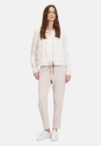 Strukturierte weiße Jacke mit rosa Zickzackmuster, gerippter weißer Oberteil und beigen Hosen mit Kordelzug. Tragen mit weißen Sneakers.