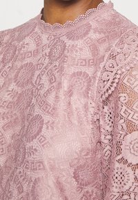 Top in pizzo rosa chiaro con intricati motivi floreali, scollatura ondulata e maniche trasparenti. Tessuto strutturato con una finitura delicata.