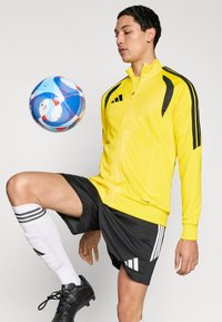 Κίτρινο αθλητικό μπουφάν με φερμουάρ και μαύρες λεπτομέρειες, σε συνδυασμό με μαύρο σορτς και λευκές κάλτσες μέχρι το γόνατο. Μια πολύχρωμη μπάλα ποδοσφαίρου αιωρείται κοντά.