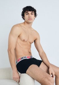 Caleçons boxeurs noirs avec une ceinture logo Diesel blanche et rouge. Le modèle a une silhouette tonique et des cheveux courts et bouclés, assis sur une surface de couleur claire.