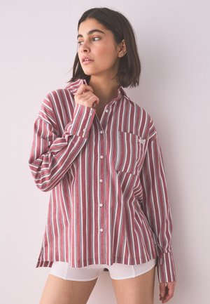 Ung kvinde med kort mørkt hår iført en oversized vinrød og hvidstribet skjorte med knapper og hvide shorts, der kigger til siden.