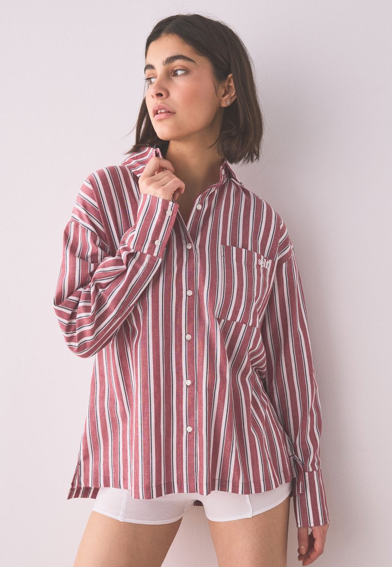 Joven con cabello corto y oscuro, vistiendo una camisa de botones de rayas grandes en burdeos y blanco, y pantalones cortos blancos, mirando hacia un lado.