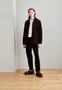 Jachetă din corduroy negru cu buzunare aplicate, combinată cu pantaloni din corduroy negru asortati și pantofi masivi negri. Cămașă albă simplă cu guler rotund dedesubt.