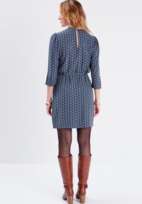 Vestido de estampado floral con mangas 3/4, cintura ajustada y detalle de escote en forma de llave en la parte trasera. Se lleva con botas marrones hasta la rodilla y medias negras.