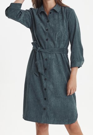 Sukienka koszulowa w ciemnym odcieniu teal, wykonana z miękkiego materiału. Posiada zapięcie na guziki z przodu, kołnierzyk i pasek w talii. Długie rękawy z podwiniętymi mankietami, lekko rozszerzany dół.