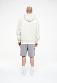 Bílá mikina s kapucí, s volným střihem a ribbovanými manžetami. Spárovaná se šedými cargo šortkami a bílými teniskami, stojící na jednobarevném pozadí.