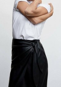 Jupe portefeuille en cuir noir avec un accent noué à la taille, dotée d'une texture lisse et d'une silhouette moderne, associée à une chemise blanche unie.