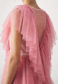 Robe en tulle rose avec des manches à volants superposés, un dos transparent et une coupe décontractée. La taille est accentuée par une ceinture en ruban assortie.