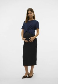 Marineblauwe ribgebreide top met geknoopte voorkant, gecombineerd met een zwarte strakke zwangerschapsrok en donkerbruine puntige slingback schoenen met studs.