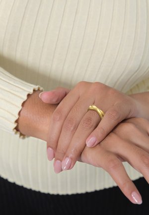 Goldenes, verdrehtes Ring auf den Fingern mit zartrosa Nagellack, Hände ruhend auf einem gerippten cremefarbenen Pullover mit gewellten Manschetten.