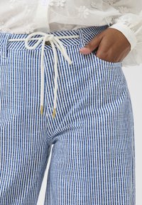Modré a bílé pruhované kalhoty s elastickým pasem, zlatě zbarvenými konci a přední kapsou, vyrobené z lehké látky.