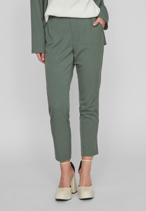 Femme portant un pantalon vert sur mesure longueur cheville, un pull blanc en côtes et des talons compensés crème avec brides à la cheville.