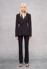 Abito nero su misura con giacca strutturata dotata di rever a punta, chiusura con un solo bottone e pantaloni affusolati coordinati. Sandali con cinturino sottile.