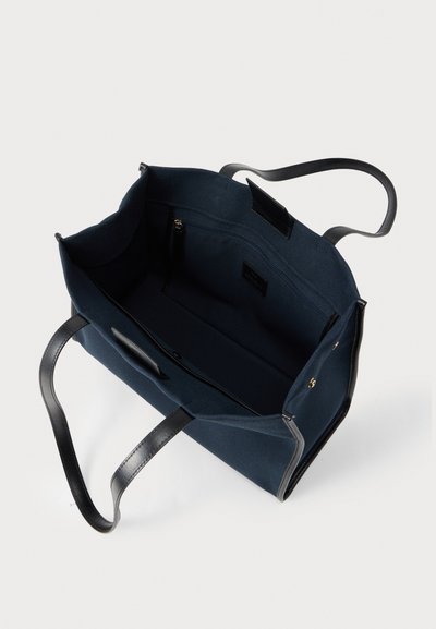 Τσάντα ώμου σε ναυτικό μπλε χρώμα με μαύρες δερμάτινες λαβές, ανοιχτό πάνω μέρος και εσωτερική τσέπη με φερμουάρ σε λευκό φόντο.