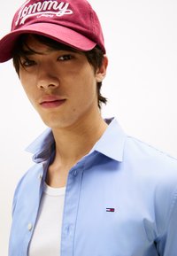 Lyseblå skjorte med knapper og hvit undertrøye. Burgunderfarget caps med hvit "Tommy"-tekst. Fokus på kragen og logodetaljen.