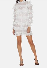 Robe blanche transparente avec des couches en volants, ornée d'accents en dentelle et en crochet, à manches longues, col haut et design ajusté.