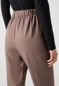 Kalhoty s elastickým pasem a rovnými nohavicemi v světle hnědé tkanině. Hladká textura, minimální detaily, model má na sobě černý top s dlouhým rukávem.
