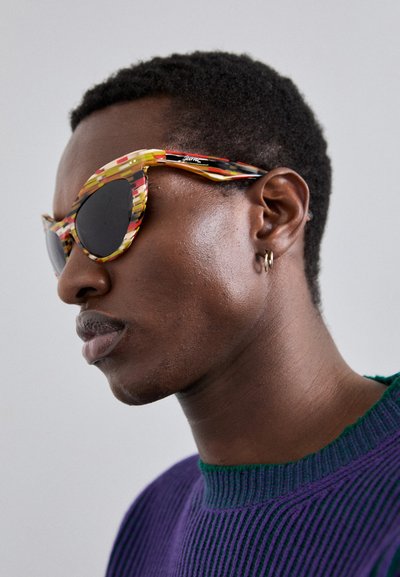 Gafas de sol multicolores con un diseño angular distintivo, que presentan patrones audaces en amarillo, rojo y negro, con lentes oscuros.