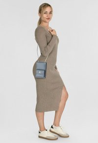 Vestito beige a maniche lunghe con motivo a coste e spacco laterale, abbinato a una piccola borsa a tracolla blu e scarpe da ginnastica bianche con suola beige.