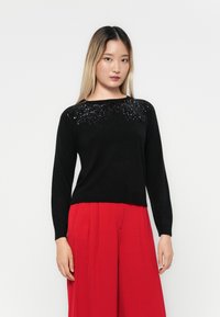 Sort sweater med lange ærmer, der har et struktureret paliet-design på den øverste front, parret med røde bredbenede bukser.