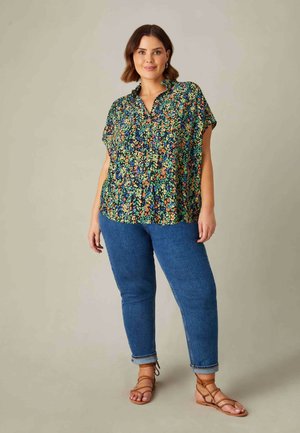 Camicia a stampa floreale nera, verde e arancione, con colletto e vestibilità rilassata, abbinata a jeans blu e sandali marroni con cinturini.