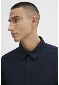 Tumedat sinine, nööpidega särk, millel on klassikaline krae, sile tekstuur ja kitsas disain. Nööbid on paigutatud esipaneelile.
